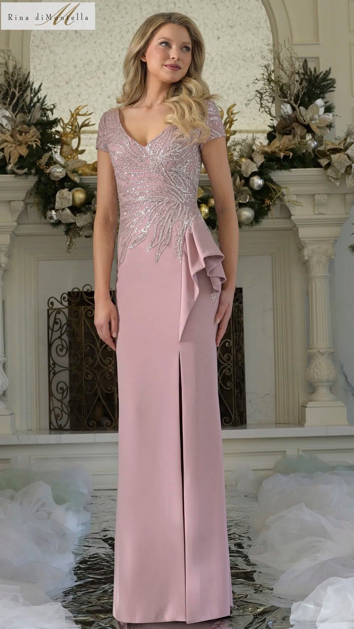 A woman in the Rina Di Montella RD3323 long formal dress in Rose, featuring sequin embellishments and a side ruffle, stands before a decorative fireplace adorned with gold and white floral arrangements.
